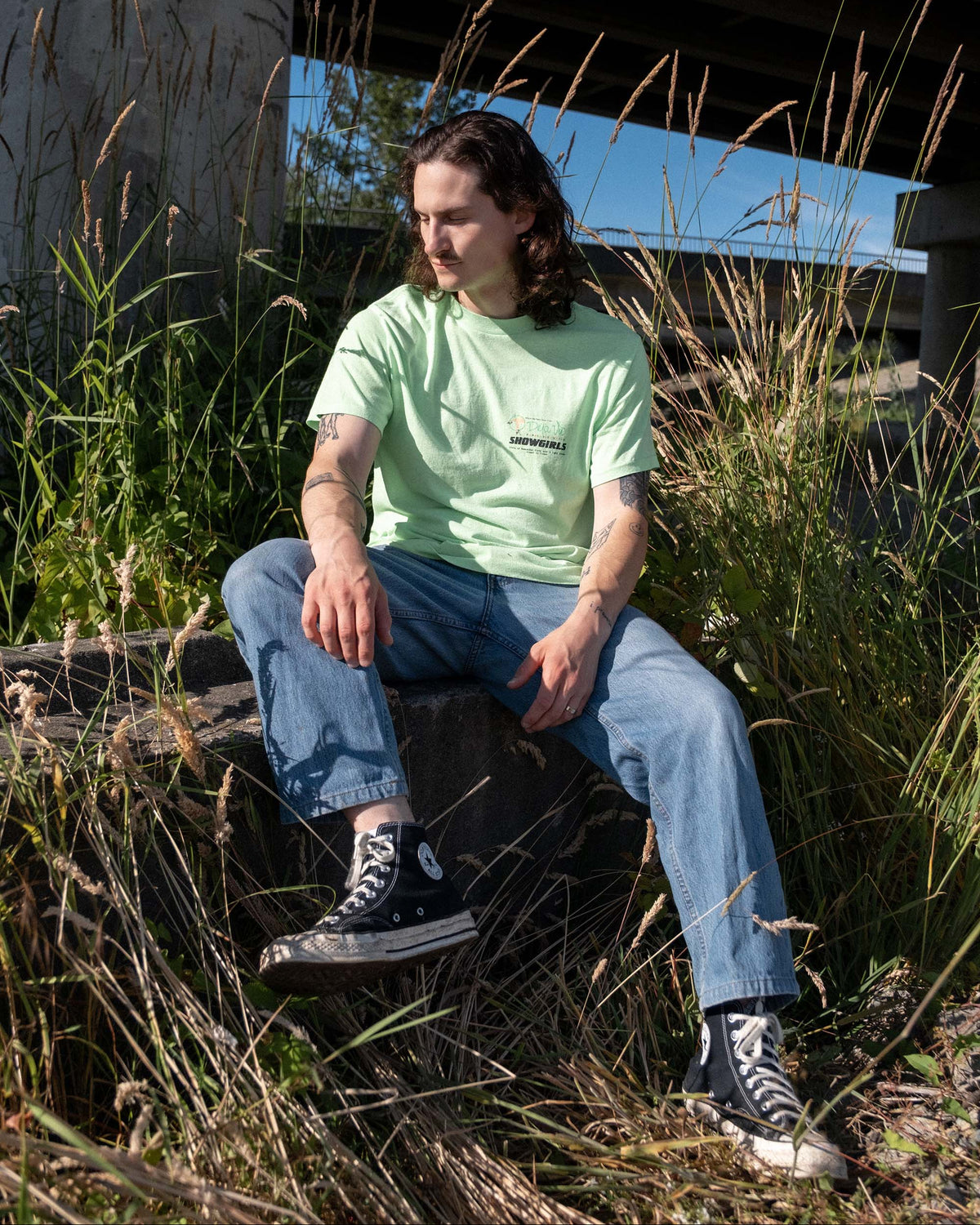 Model wearing late-1980s Deja Vu Showgirls neon green graphic T-shirt outdoors beneath an overpass.